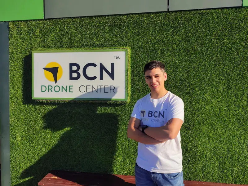 L’Adrià, amb els braços creuats i una samarreta de BCN Drone Center, davant del rètol de l’empresa en una paret de gespa artificial.